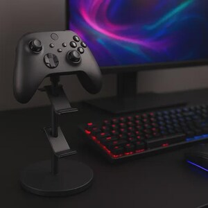 Puede incluir: Un mando de videojuegos negro sobre un soporte negro. El soporte tiene una base circular y dos plataformas anguladas. En el fondo se ve un teclado con teclas retroiluminadas en rojo y azul.
