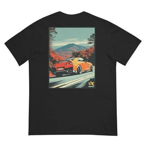 Op de afbeelding: Zwart T-shirt met een afbeelding van een oranje en rode sportwagen op een bergweg. De achtergrond toont herfstkleuren en bergen. Het shirt bevat de tekst "Rogue & Rally".