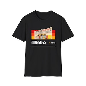 May include: A black short-sleeve t-shirt featuring a graphic print of a storefront with the text "BUILDING GOOD STUFF CHEAP" above it. Below the image, the word "Retro" is printed in white with a pixelated effect, followed by "Vibes".