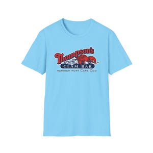 May include: Pale blue t-shirt with the text "Thompson's Clam Bar, Harwich Port Cape Cod" in red and white. The design includes a red lobster and clams. The shirt has a crew neck.