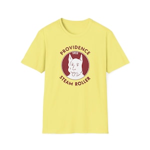 May include: A light yellow t-shirt featuring a circular graphic with a dog illustration and the text "PROVIDENCE 1922 STEAM ROLLER" in a maroon color. The shirt has short sleeves and a crew neck.