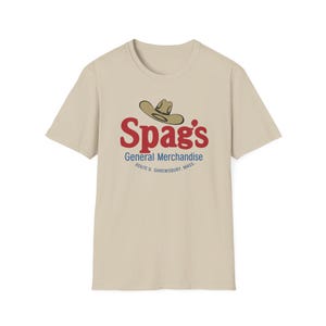 May include: A beige short-sleeve t-shirt featuring a graphic of a tan cowboy hat above the red text Spag's, followed by the blue text General Merchandise and the smaller blue text ROUTE 9, SHREWSBURY, MASS. centered on the chest.
