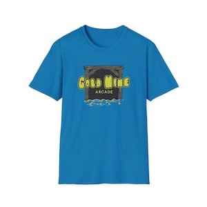 May include: A blue t-shirt with a graphic depicting a mine entrance. The words "Gold Mine Arcade" are in yellow. The graphic has a wooden frame and a dark background with yellow accents.