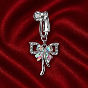 May include: A silver-colored belly button ring with a bow pendant. The bow has a shimmering, iridescent finish and is encrusted with small, clear crystals. The top of the ring features a round, crystal-studded clasp.
