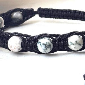 Könnte beinhalten: Ein Makramee-Armband mit weiß-grün gesprenkelten Perlen. Die Perlen sind in ein schwarzes Makramee-Design eingewebt. Das Armband hat verstellbare schwarze Kordeln mit kleinen weißen Perlen an den Enden.