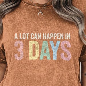 Peut inclure: T-shirt orange-brun avec le texte "A LOT CAN HAPPEN IN 3 DAYS" en couleurs pastel. Le t-shirt a un aspect vintage et délavé, avec des manches retroussées. Le modèle porte un short en jean.