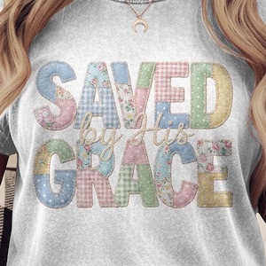 May include: A light gray t-shirt with the text "SAVED by His GRACE" in a patchwork design of pastel colors. The shirt has rolled-up sleeves and a crew neck. The model is wearing light-wash denim jeans.