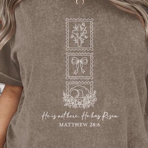 Könnte beinhalten: Braunes T-Shirt mit weißen grafischen Briefmarken und dem Text "He is not here. He has Risen. Matthew 28:6." Die Briefmarken zeigen ein Kreuz, eine Schleife und ein Blumendesign. Die Ärmel sind hochgekrempelt.