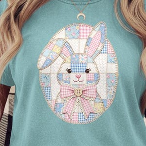 Peut inclure: Un t-shirt bleu clair avec un motif de lapin de Pâques en patchwork. Le lapin est à l'intérieur d'une forme ovale et présente des accents roses, bleus et jaunes. Le lapin a un nœud rose et un bouton. Le t-shirt a des manches retroussées.