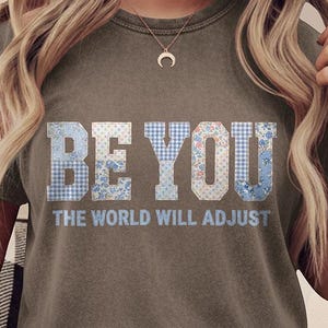 May include: Olive green t-shirt with the text "BE YOU" in a patchwork design, and the phrase "THE WORLD WILL ADJUST" below. The shirt has rolled-up sleeves and is worn with light-wash jeans.