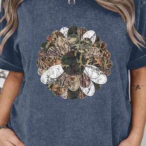 May include: A navy blue t-shirt with a large floral design. The flower's petals are a mix of camouflage patterns and white, with a camouflage centre. The shirt has short sleeves and a relaxed fit.