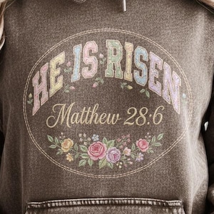 May include: Brown hooded sweatshirt with a front pocket and drawstring. The front features a circular design with the text "HE IS RISEN" in pastel colors, along with "Matthew 28:6" and floral accents. The sweatshirt has a vintage wash.