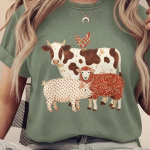 May include: Sage green t-shirt featuring a whimsical illustration of farm animals. A cow with brown spots, a chicken, a pig with polka dots, and a paisley-patterned sheep are depicted. The shirt has short sleeves and a crew neck.