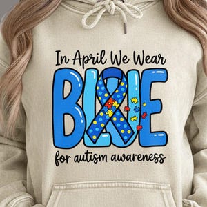 以下が含まれることがあります： 「In April We Wear BLUE for autism awareness」と書かれた青いデザインのベージュ色のパーカー。「BLUE」の文字はリボンとパズルのピースで装飾されています。