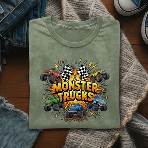 Pu&ograve; includere: Una maglietta verde salvia con una grafica colorata di monster truck. Il design include monster truck arancioni, gialli, verdi e blu, bandiere a scacchi e le parole "MONSTER TRUCKS" in grassetto, arancione.
