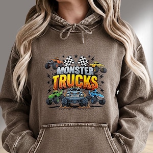 K&ouml;nnte beinhalten: Brauner Kapuzenpullover mit einem Front-Grafik-Design mit dem Text "MONSTER TRUCKS" und Illustrationen von Monster Trucks in verschiedenen Farben. Das Design umfasst karierte Flaggen und Sterne.