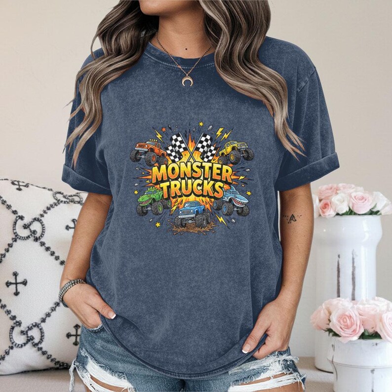 Pu&ograve; includere: T-shirt blu navy con una grafica colorata di monster truck. Il design include il testo "MONSTER TRUCKS" con bandiere a scacchi e illustrazioni di monster truck arancioni, verdi, gialli e blu.