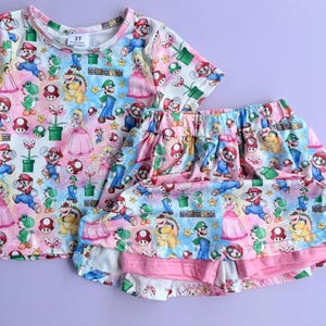 May include: A two-piece children's outfit featuring a colorful print of characters from the Super Mario video game series. The set includes a short-sleeved top and a skirt with a ruffled hem. The outfit is size 3T.