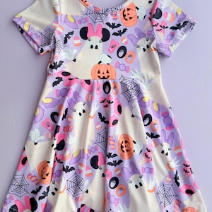 May include: A short-sleeve dress with a light purple background featuring a repeating Halloween pattern of ghosts wearing pink bows, orange pumpkins, black spiderwebs, bats, and colorful candy corn. The dress has a round neckline and a flared skirt.