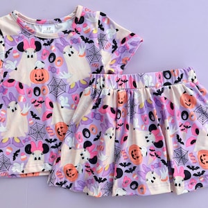 May include: A short-sleeve t-shirt and a pleated skirt set featuring a repeating Halloween pattern on a light purple background. The print includes white ghost figures with pink bows, orange jack-o'-lanterns, black spiderwebs, bats, and candy. The shirt tag reads 3T.