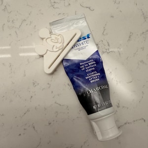 Puede incluir: Un tubo de pasta dental Crest Whitening Charcoal con un exprimidor de tubo de pasta dental blanco con forma de Mickey Mouse. El tubo de pasta dental es azul y blanco, con el texto "Removes up to 90% of surface stains". El tubo contiene 108g de pasta dental.