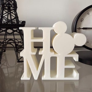 May include: A cream-colored decorative sign that spells out "HOME". The "O" is replaced with a Mickey Mouse head silhouette. A black Eiffel Tower and a clock are visible in the background.