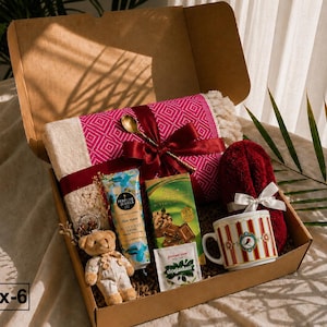K&ouml;nnte beinhalten: Eine Geschenkschachtel aus Karton mit einer rosa-cremefarbenen gemusterten Decke, die mit einem kastanienbraunen Band und einem goldenen L&ouml;ffel gebunden ist, einem kleinen Teddyb&auml;ren, Handcreme, Schokolade, Teebeutel und einer gestreiften Tasse.