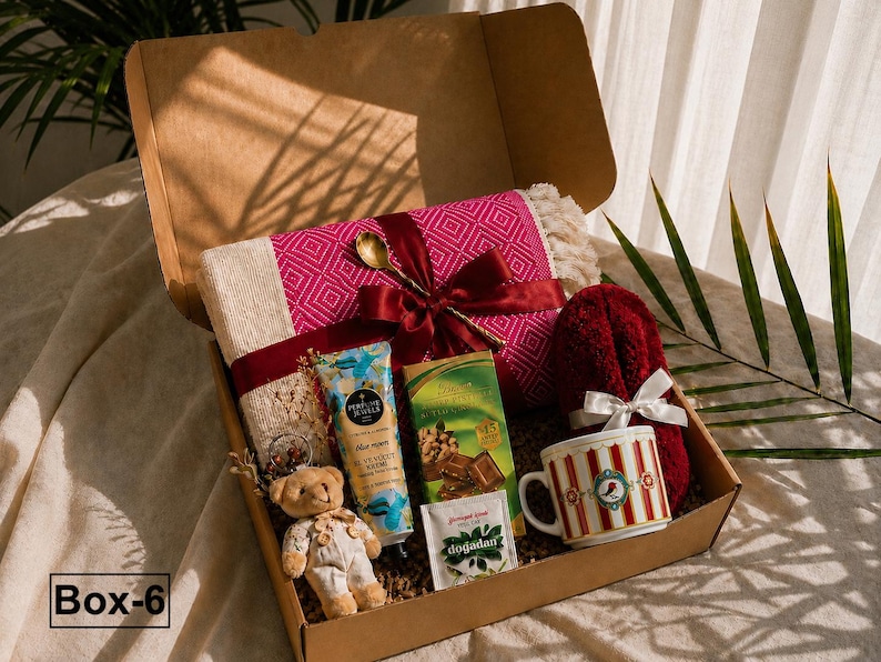 K&ouml;nnte beinhalten: Eine Geschenkschachtel aus Karton mit einer rosa-cremefarbenen gemusterten Decke, die mit einem kastanienbraunen Band und einem goldenen L&ouml;ffel gebunden ist, einem kleinen Teddyb&auml;ren, Handcreme, Schokolade, Teebeutel und einer gestreiften Tasse.