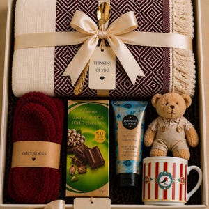 May include: A gift box containing a burgundy and cream diamond-patterned throw blanket with fringe, a pair of burgundy socks, a bar of pistachio milk chocolate, a 75ml tube of hand cream, a small teddy bear, and a striped mug with a bird graphic.
