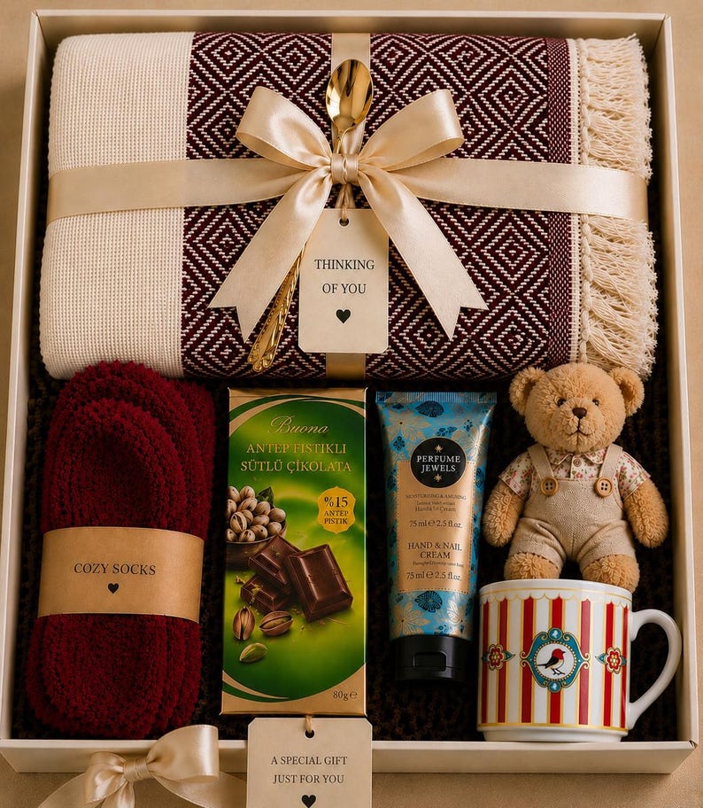 May include: A gift box containing a burgundy and cream diamond-patterned throw blanket with fringe, a pair of burgundy socks, a bar of pistachio milk chocolate, a 75ml tube of hand cream, a small teddy bear, and a striped mug with a bird graphic.