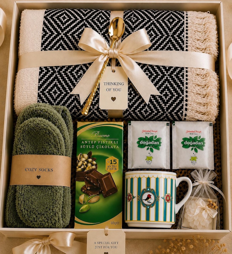 May include: A gift box containing a black and white patterned throw blanket with fringe, a gold spoon, a tag reading Thinking of You, olive green fuzzy socks, a bar of Buono chocolate, two Dogadan tea bags, a striped mug, and a small bag of white flowers.