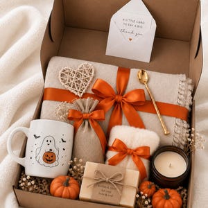 May include: A gift box containing a white mug with a ghost and pumpkin print, a beige blanket tied with an orange ribbon, a small burlap pouch, a bar of soap labeled Natural Soap, a candle, a gold spoon, a heart ornament, and a thank you card.