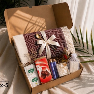 K&ouml;nnte beinhalten: Eine Geschenkschachtel aus Karton mit einer gefalteten, bordeaux-wei&szlig; gemusterten Decke mit Fransen, die mit einem cremefarbenen Band und einem goldenen L&ouml;ffel verziert ist. Enth&auml;lt zwei Teep&auml;ckchen, eine Schokolade, zwei Glasphiolen, Bl&uuml;ten und Seife.