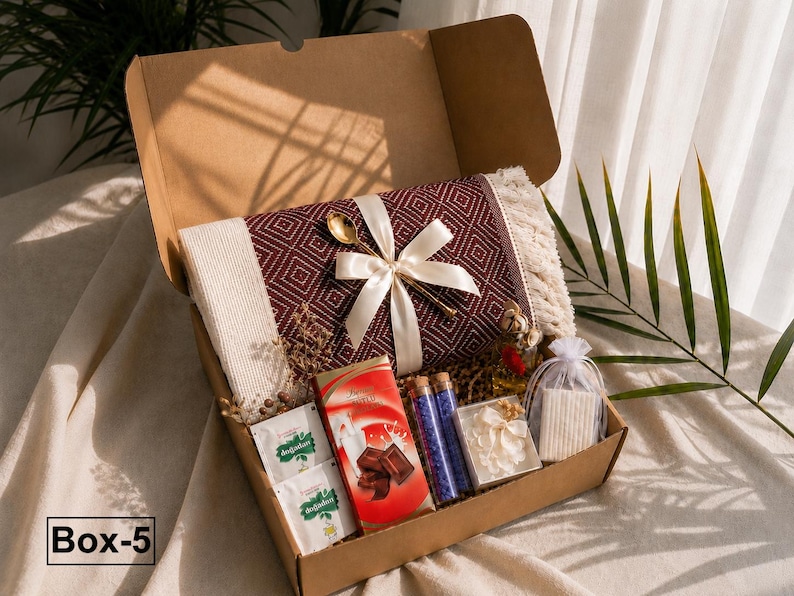 K&ouml;nnte beinhalten: Eine Geschenkschachtel aus Karton mit einer gefalteten, bordeaux-wei&szlig; gemusterten Decke mit Fransen, die mit einem cremefarbenen Band und einem goldenen L&ouml;ffel verziert ist. Enth&auml;lt zwei Teep&auml;ckchen, eine Schokolade, zwei Glasphiolen, Bl&uuml;ten und Seife.
