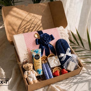 K&ouml;nnte beinhalten: Eine mit Box-7 beschriftete Kartonschachtel enth&auml;lt eine rosa-wei&szlig; gemusterte Decke mit dunkelblauem Band und Blume, einen kleinen Teddyb&auml;ren, eine Tube Perfume Jewels Handcreme, zwei Glasfl&auml;schchen mit violettem Badesalz und ein dunkelblaues Handtuch.
