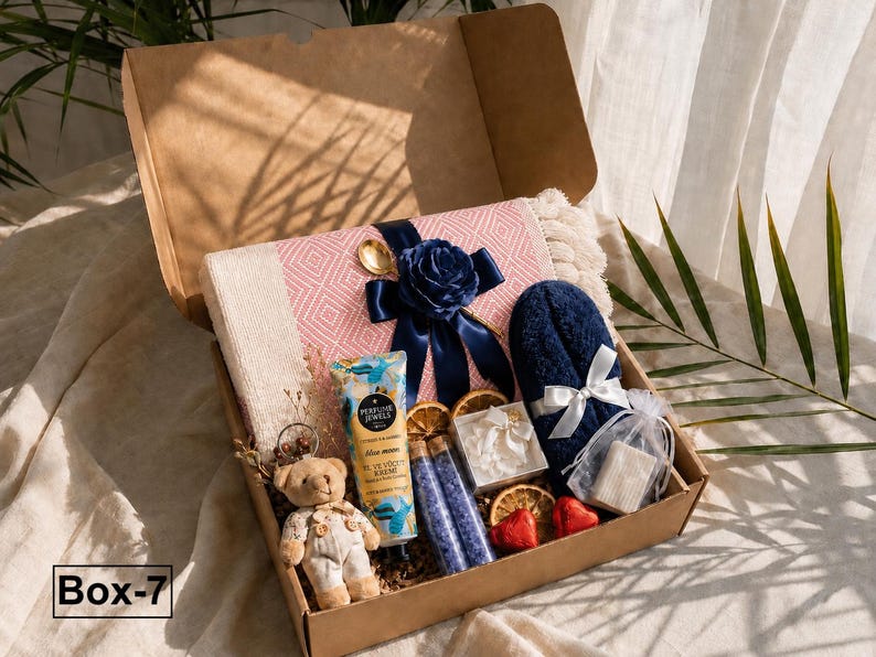 K&ouml;nnte beinhalten: Eine mit Box-7 beschriftete Kartonschachtel enth&auml;lt eine rosa-wei&szlig; gemusterte Decke mit dunkelblauem Band und Blume, einen kleinen Teddyb&auml;ren, eine Tube Perfume Jewels Handcreme, zwei Glasfl&auml;schchen mit violettem Badesalz und ein dunkelblaues Handtuch.