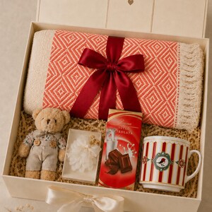 May include: A gift box containing a folded coral and cream diamond-patterned throw blanket with fringe tied with a dark red ribbon, a small plush teddy bear, a white floral ornament, a bar of Buena S&uuml;tl&uuml; &Ccedil;ikolata, and a red and white striped mug.