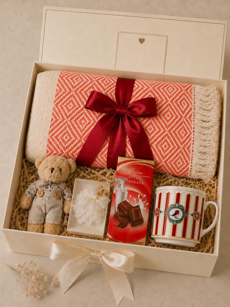 May include: A gift box containing a folded coral and cream diamond-patterned throw blanket with fringe tied with a dark red ribbon, a small plush teddy bear, a white floral ornament, a bar of Buena S&uuml;tl&uuml; &Ccedil;ikolata, and a red and white striped mug.