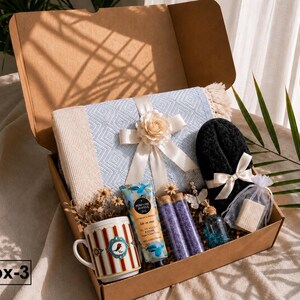 K&ouml;nnte beinhalten: Eine Geschenkbox mit einer hellblauen, gemusterten Decke mit cremefarbener Schleife und Blume, einem wei&szlig;en Becher mit roten Streifen und Vogelmotiv, einer Handcreme, zwei violetten Badesalzr&ouml;hrchen, einem kleinen Glasfl&auml;schchen und schwarzen Hausschuhen.