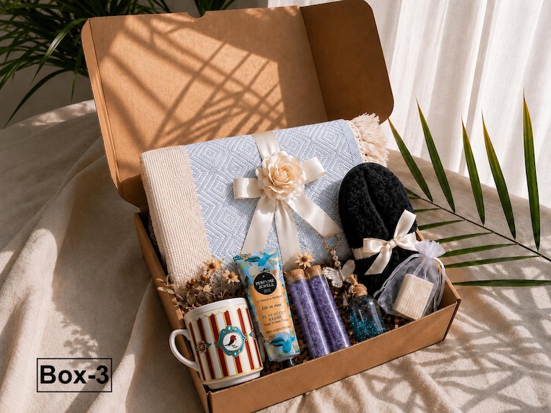 K&ouml;nnte beinhalten: Eine Geschenkbox mit einer hellblauen, gemusterten Decke mit cremefarbener Schleife und Blume, einem wei&szlig;en Becher mit roten Streifen und Vogelmotiv, einer Handcreme, zwei violetten Badesalzr&ouml;hrchen, einem kleinen Glasfl&auml;schchen und schwarzen Hausschuhen.
