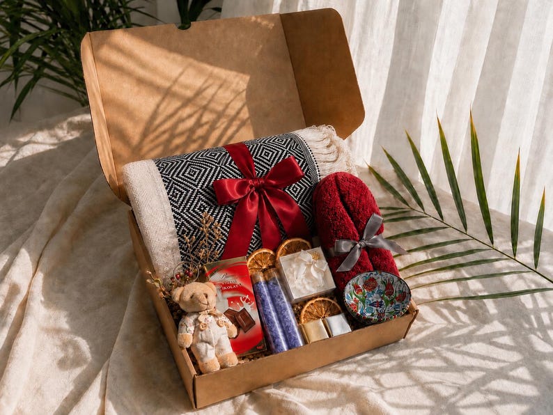 K&ouml;nnte beinhalten: Eine Geschenkschachtel aus Karton mit einer schwarz-wei&szlig; gemusterten Wohndecke mit roter Schleife, einem roten Handtuch mit grauer Schleife, einem kleinen Teddyb&auml;ren, einer Schokolade, zwei Glasfl&auml;schchen mit violettem Badesalz und einer Blumenschale.