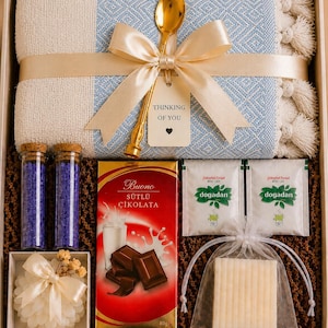 May include: A gift box containing a light blue and cream woven textile with tassels, a gold spoon, a tag reading Thinking of You, two glass vials of purple bath salts, a bar of Buono milk chocolate, two Dogadan tea bags, and a white soap bar.