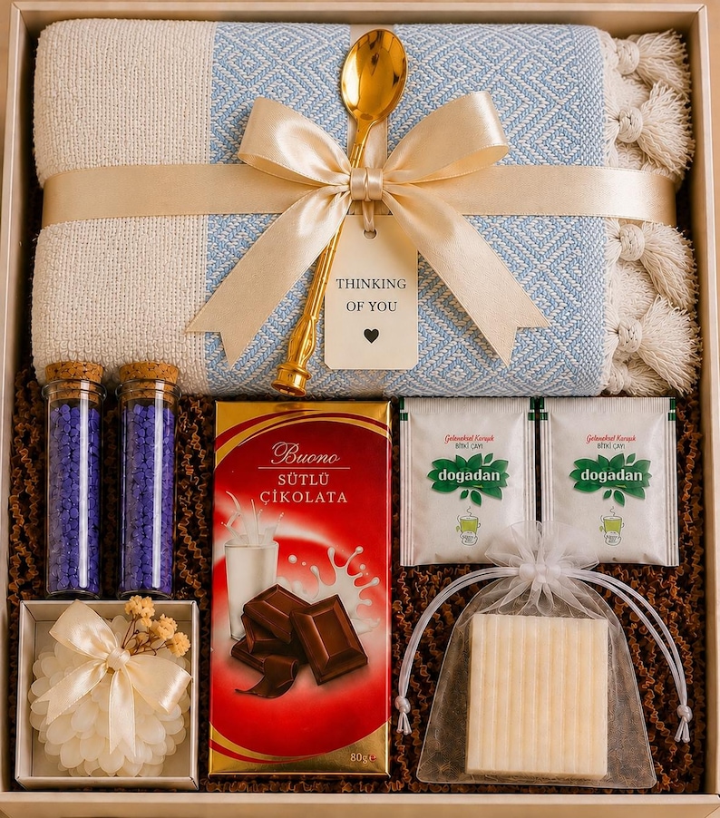May include: A gift box containing a light blue and cream woven textile with tassels, a gold spoon, a tag reading Thinking of You, two glass vials of purple bath salts, a bar of Buono milk chocolate, two Dogadan tea bags, and a white soap bar.