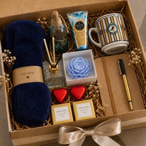 May include: A gift box containing navy blue fuzzy socks with a label reading COZY SOCKS, a glass reed diffuser, a blue flower-shaped candle, a tube of hand cream, a striped mug with a bird graphic, a notebook, a pen, and two chocolate squares.