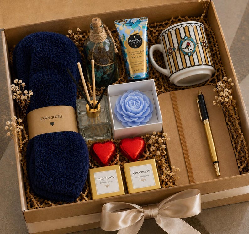 May include: A gift box containing navy blue fuzzy socks with a label reading COZY SOCKS, a glass reed diffuser, a blue flower-shaped candle, a tube of hand cream, a striped mug with a bird graphic, a notebook, a pen, and two chocolate squares.