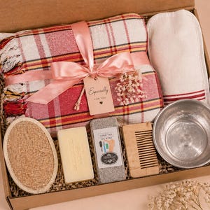 May include: A gift box containing a red and white plaid towel with a pink ribbon and a tag reading Especially for you, a white mitt, a round textured sponge, a rectangular soap bar, a gray pumice stone, a wooden comb, and a metal bowl.