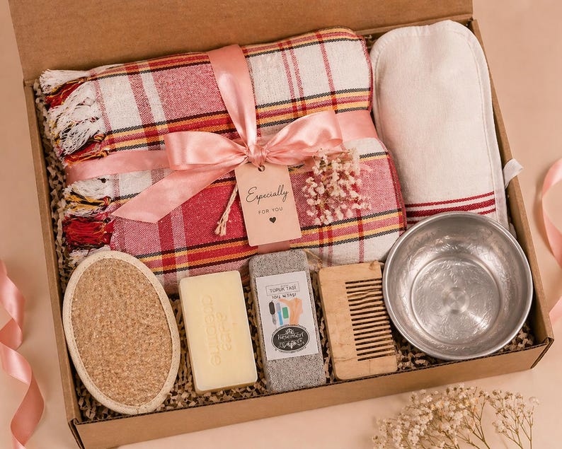 May include: A gift box containing a red and white plaid towel with a pink ribbon and a tag reading Especially for you, a white mitt, a round textured sponge, a rectangular soap bar, a gray pumice stone, a wooden comb, and a metal bowl.
