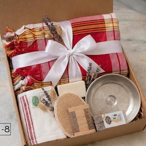 K&ouml;nnte beinhalten: Eine Geschenkbox mit der Aufschrift Box-8, die ein rot-gelb kariertes Textil mit wei&szlig;em Band, ein rechteckiges Seifenst&uuml;ck, einen runden Luffaschwamm, einen Holzkamm, eine kleine Metallschale mit Blattmustern und getrocknete Lavendelzweige enth&auml;lt.