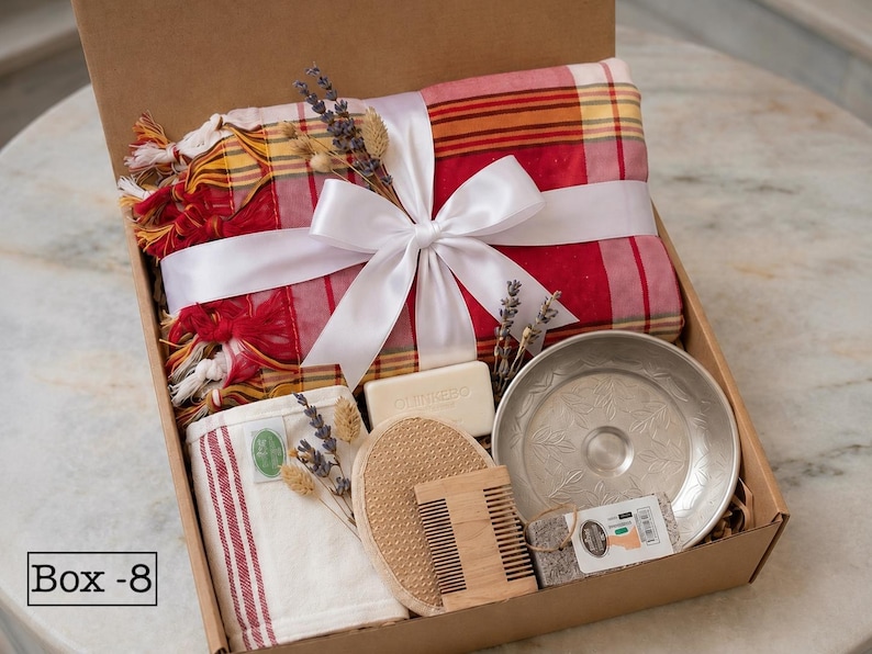 K&ouml;nnte beinhalten: Eine Geschenkbox mit der Aufschrift Box-8, die ein rot-gelb kariertes Textil mit wei&szlig;em Band, ein rechteckiges Seifenst&uuml;ck, einen runden Luffaschwamm, einen Holzkamm, eine kleine Metallschale mit Blattmustern und getrocknete Lavendelzweige enth&auml;lt.