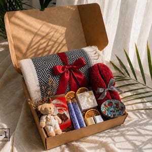 K&ouml;nnte beinhalten: Eine Geschenkschachtel aus Karton mit einer schwarz-wei&szlig; gemusterten Decke mit roter Schleife, einem kleinen Teddyb&auml;ren, einer roten Schokoladentafel, zwei violetten Badesalzr&ouml;hrchen, getrockneten Orangenscheiben und einem roten Handtuch.